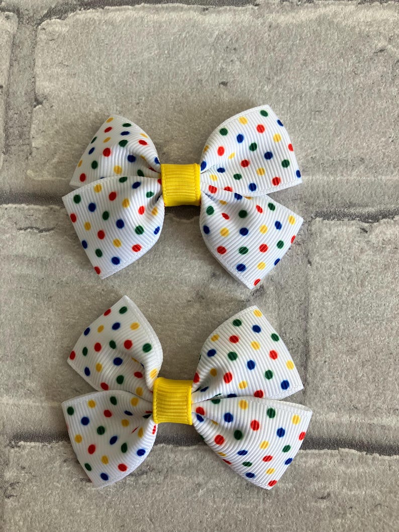 May include: Two white hair bows with a yellow center and multi-colored polka dots. The polka dots are red, blue, green, and yellow.