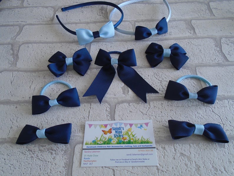 May include: An assortment of navy blue and light blue hair accessories, including headbands, bows, and hair ties. The bows have a contrasting light blue center. A business card with the text "Sandi's Mini Styles" is also visible.
