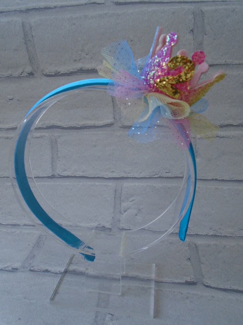Puede incluir: Una diadema turquesa con un lazo de tul rosa, amarillo y azul y una corona de purpurina.