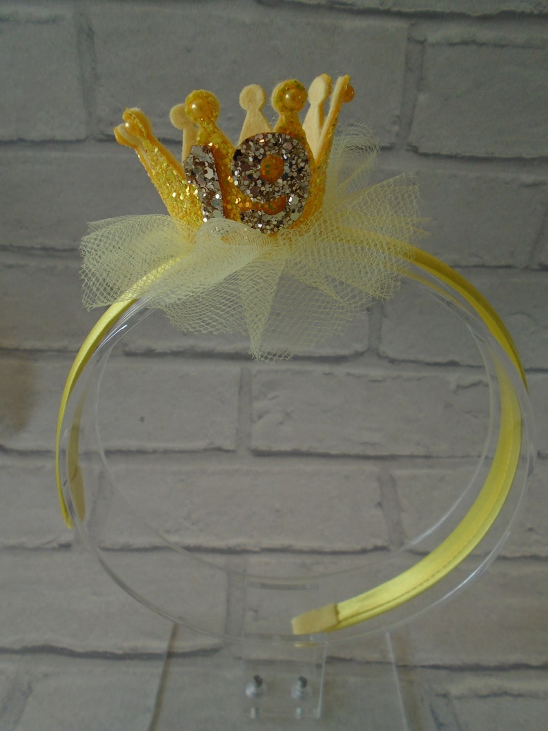 Puede incluir: Diadema de brillantina amarilla con una corona amarilla y el n&uacute;mero 19 en ella. La corona est&aacute; decorada con diamantes de imitaci&oacute;n.