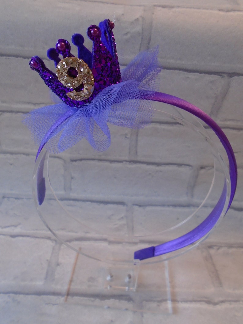 Puede incluir: Diadema morada con una corona morada brillante con un n&uacute;mero nueve de purpurina dorada. La corona tiene un lazo de tul azul claro.