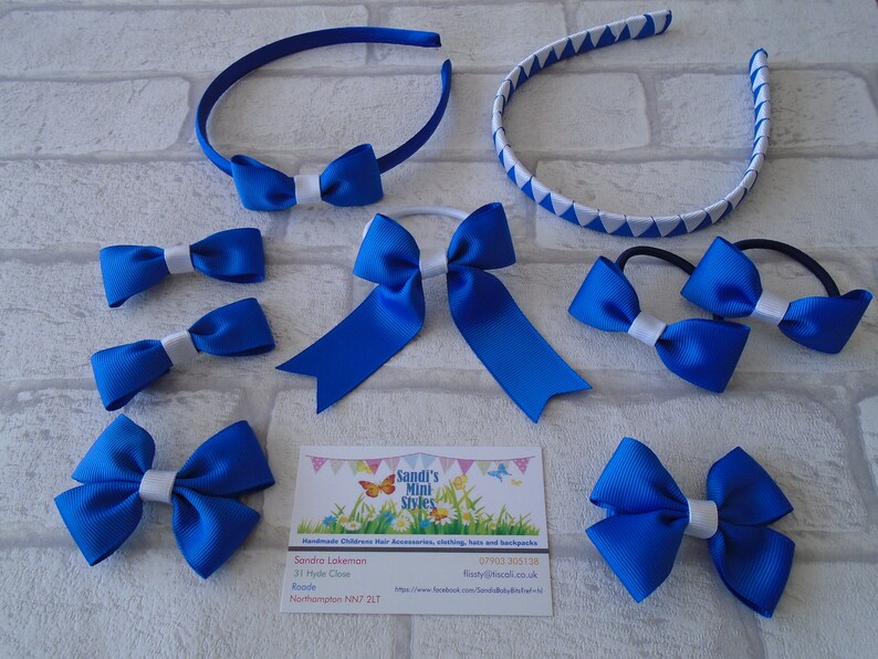 May include: An assortment of blue hair accessories, including headbands, bow ties, and hair ties. Some bows have a white center. A business card with the text "Sandi's Mini Styles" is also present.