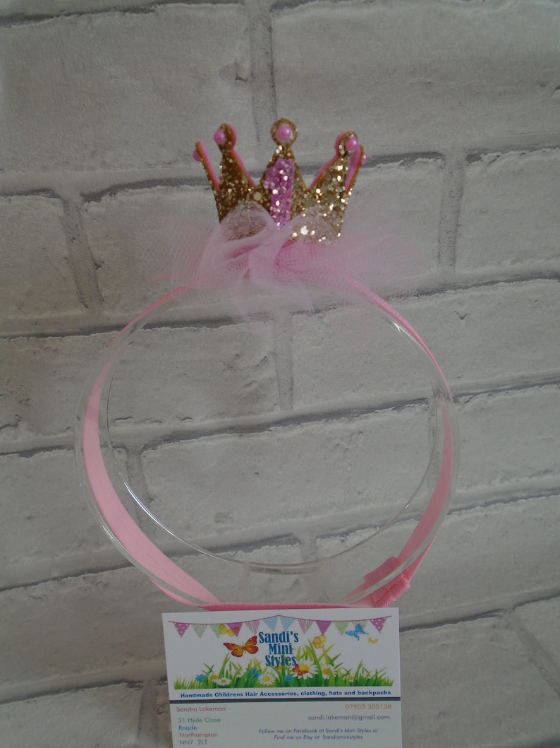 Puede incluir: Diadema con corona de purpurina rosa y dorada con un lazo de tul. La diadema est&aacute; sobre un fondo de ladrillo blanco. La tarjeta de visita dice "Sandi's Mini Styles Accesorios para el cabello, ropa, sombreros y mochilas para ni&ntilde;os hechos a mano".