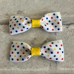May include: Two white bow hair clips with a yellow center and a polka dot pattern in red, blue, green, and yellow.