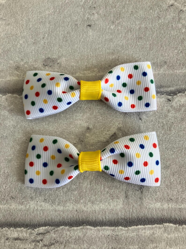 May include: Two white bow hair clips with a yellow center and a polka dot pattern in red, blue, green, and yellow.