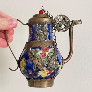 Puede incluir: Una tetera pequeña azul y dorada con un diseño de dragón. La tetera tiene un asa y un pico de metal, y una cadena con un amuleto de dragón decorativo que cuelga de la tapa.