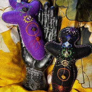 May include: Two felt dolls in the shape of a person, one purple and one black, with embroidered details and gemstone accents. The dolls are displayed on a black hand sculpture against a yellow and green patterned background.