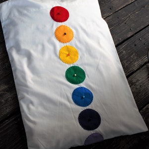 May include: A white pillow with a row of seven colorful felt circles sewn on, each with a small button in the center. The circles are red, orange, yellow, green, blue, indigo, and violet.
