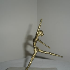 May include: A gold-colored metal sculpture of a dancer in mid-pose. The dancer is in a graceful, flowing position with one leg extended and the other bent. The sculpture is a beautiful example of the art of dance.
