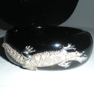 May include: Black bangle bracelet with a silver lizard design. The lizard is adorned with rhinestones.