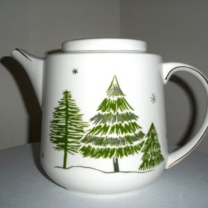 May include: White ceramic teapot with a lid, featuring green Christmas tree illustrations and gold trim on the handle. The teapot has a spout and a handle. Snowflakes are also depicted on the teapot. A winter holiday theme.