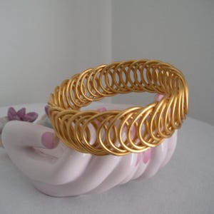 May include: A gold-colored metal bracelet with a woven design, resting on a white and pink decorative hand-shaped dish. The bracelet's design features a series of connected loops.