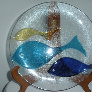 Puede incluir: Un plato de vidrio transparente con una superficie texturizada presenta tres peces, uno amarillo, uno azul y uno azul oscuro. Los peces están hechos de vidrio y tienen un acabado brillante.
