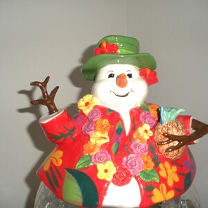May include: A ceramic snowman figurine with a white face, orange carrot nose, and a green hat adorned with red flowers. The snowman wears a red Hawaiian shirt decorated with colorful flowers and holds a pineapple.