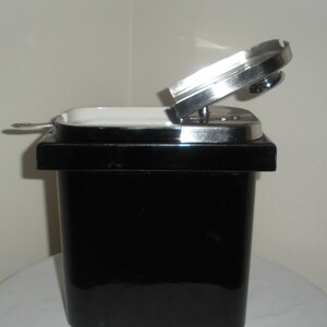 May include: A black ceramic container with a white interior and a silver lid. The lid is open and the container is sitting on a white surface.