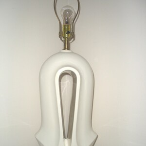May include: A white ceramic table lamp with a unique, arched design. The lamp features a gold-colored base and a brass neck supporting a light bulb. The lamp's height is approximately 20 inches.