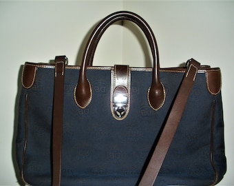 May include: A dark blue canvas tote bag with brown leather trim and handles. The bag has a top zipper closure and a long shoulder strap. The bag is embossed with a repeating pattern.