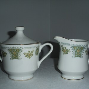 May include: A white porcelain sugar bowl with a lid and a creamer. Both pieces feature a decorative floral pattern in green and yellow, with gold trim. The sugar bowl has two handles.