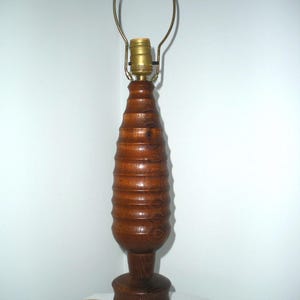 May include: A vintage wooden table lamp with a carved, textured design. The lamp has a bulb socket and a brass-colored harp. The base is round and the lamp is approximately 24 inches tall.