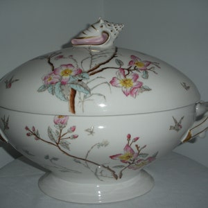 May include: A white porcelain serving dish with a lid. The dish is decorated with a floral pattern of pink and yellow flowers and green leaves. The lid has a seashell finial.