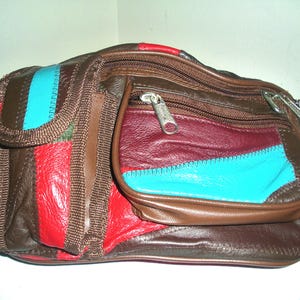 May include: A brown leather fanny pack with a patchwork design. The bag features red, teal, and burgundy panels, with a zippered main compartment and a smaller front pocket. The bag has a brown strap and silver zippers.