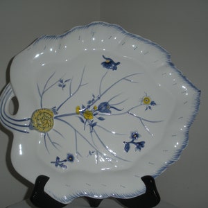 Puede incluir: Un plato de servir de cerámica blanca con forma de hoja con diseños florales azules y amarillos. El plato tiene dos asas y un diseño de caracol en relieve.