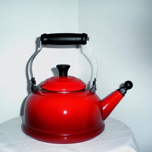 May include: A shiny red tea kettle with a black handle and spout. The kettle has a black lid and a silver handle that arches over the top. The kettle is sitting on a white surface.
