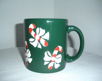 May include: A green ceramic mug with a white and red candy cane design. The mug has a white bow on each candy cane.