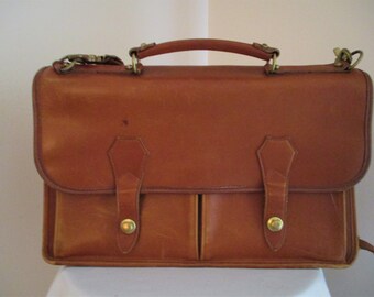 May include: A brown leather briefcase with a top handle and a shoulder strap. The briefcase has two front pockets with brass buckles and two brass buttons.