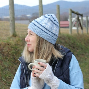 May include: A woman wearing a blue and white knit hat and a blue vest holds a white mug with a blue and white design. She is wearing a blue long-sleeved shirt and white fingerless gloves.