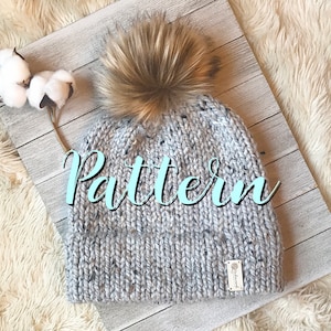 May include: A light gray knit beanie with a faux fur pom-pom. The hat has a speckled design and a small tag. The word "Pattern" is written across the front of the hat in a cursive font. The hat is displayed on a white wooden surface.