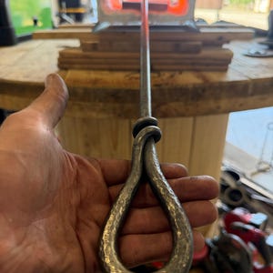 Op de afbeelding: Een handgesmede ijzeren pook met een lange, rechte schacht en een lusvormig handvat met een getextureerde, gehamerde afwerking. De metalen staaf strekt zich uit naar een gloeiende, oranje-hete smidse op de achtergrond.