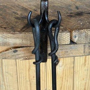 May include: A black metal wall-mounted rack featuring two curved hooks extending from a central vertical plate. The dark, textured iron surface shows visible hammer marks, with two long, slender rods hanging from the hooks by looped ends.