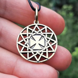 May include: Gold-colored pendant with a geometric design, featuring a central cross within a square and surrounded by a starburst pattern. The pendant hangs from a black cord. The background is blurred green foliage.