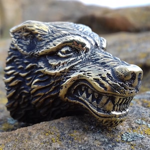 Op de afbeelding: Een gedetailleerde, goudkleurige metalen ring met een wolvenkop met een grommende uitdrukking. De vacht van de wolf is getextureerd en de ring ligt op een rotsachtige achtergrond.