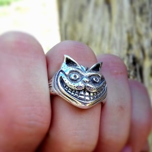 Puede incluir: Un anillo de plata con el Gato de Cheshire de Alicia en el país de las maravillas. El anillo muestra la sonrisa y los rasgos faciales icónicos del gato. El anillo se lleva en un dedo, con el fondo borroso.