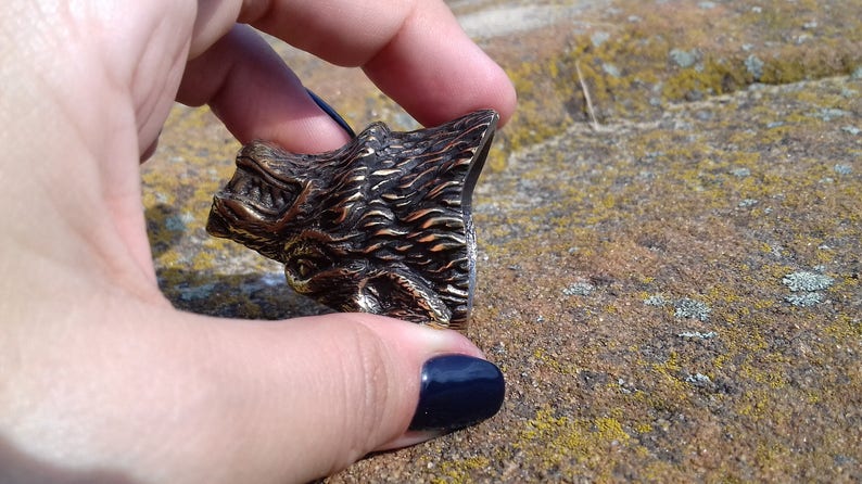 May include: A detailed bronze wolf head sculpture with a textured fur design. The sculpture is held in a hand with a dark blue nail polish.