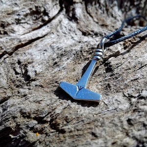 May include: A silver-colored pendant in the shape of Thor's hammer, Mjolnir, hangs from a black cord. The hammer has a small engraved symbol on its head. The pendant is set against a textured, weathered wood background.