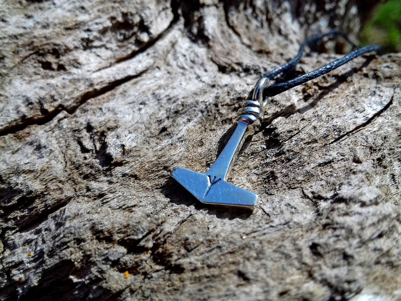 May include: A silver-colored pendant in the shape of Thor's hammer, Mjolnir, hangs from a black cord. The hammer has a small engraved symbol on its head. The pendant is set against a textured, weathered wood background.