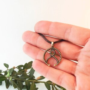 May include: A bronze pendant necklace with a circular design featuring a stylized figure with a beaded border. The pendant is hanging from a brown cord.