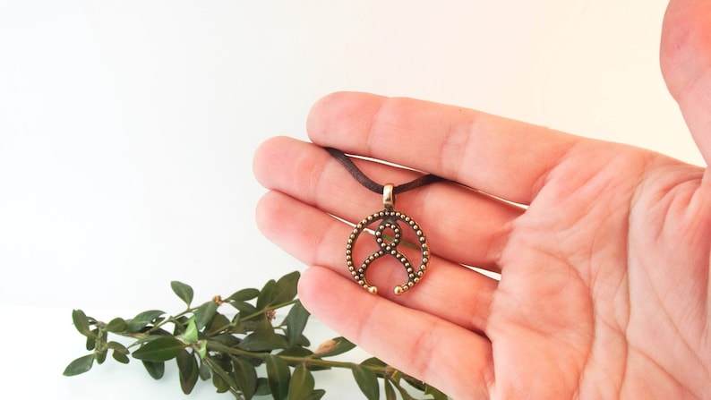 May include: A bronze pendant necklace with a circular design featuring a stylized figure with a beaded border. The pendant is hanging from a brown cord.
