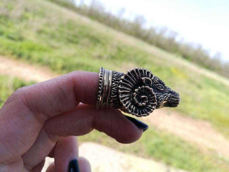 May include: A gold-toned ring featuring a detailed ram head design. The ram's horns are curled and the ring is worn on a finger.