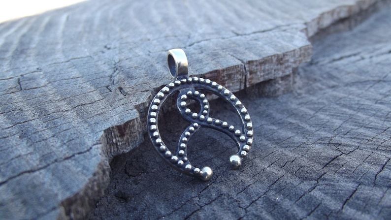 May include: A silver pendant with a circular design featuring a stylized knot or symbol. The pendant is adorned with small beads around the edges and the knot.