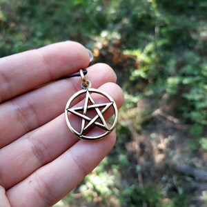 May include: A gold-colored pendant featuring a five-pointed star, also known as a pentagram, within a circle. The pendant is held in a hand against a blurred green background.