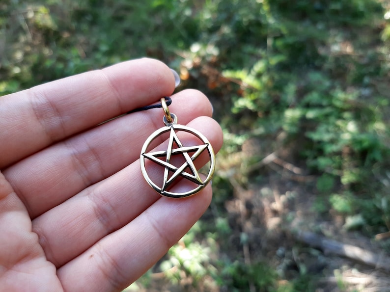 May include: A gold-colored pendant featuring a five-pointed star, also known as a pentagram, within a circle. The pendant is held in a hand against a blurred green background.