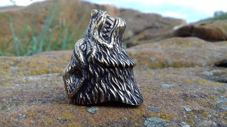 May include: A detailed bronze ring featuring a wolf's head with its mouth open, showing sharp teeth. The wolf's fur is textured and the ring is set against a rocky background.