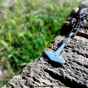 May include: A silver pendant necklace featuring a hammer design, possibly Mjolnir, with a rune symbol. The pendant hangs from a black cord, resting on a weathered wooden surface with green grass in the background.