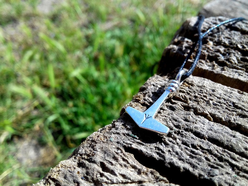 May include: A silver pendant necklace featuring a hammer design, possibly Mjolnir, with a rune symbol. The pendant hangs from a black cord, resting on a weathered wooden surface with green grass in the background.