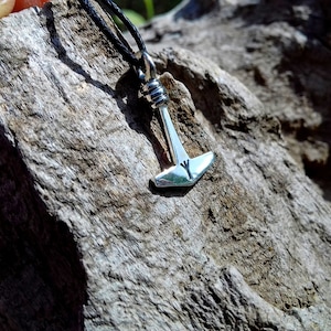 May include: A silver pendant necklace featuring a small hammer-shaped charm, likely representing Thor's hammer, Mjolnir. The pendant is suspended from a black cord and rests against a textured, gray wooden surface. The hammer has a rune symbol.