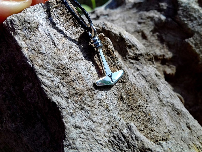 May include: A silver pendant necklace featuring a small hammer-shaped charm, likely representing Thor's hammer, Mjolnir. The pendant is suspended from a black cord and rests against a textured, gray wooden surface. The hammer has a rune symbol.